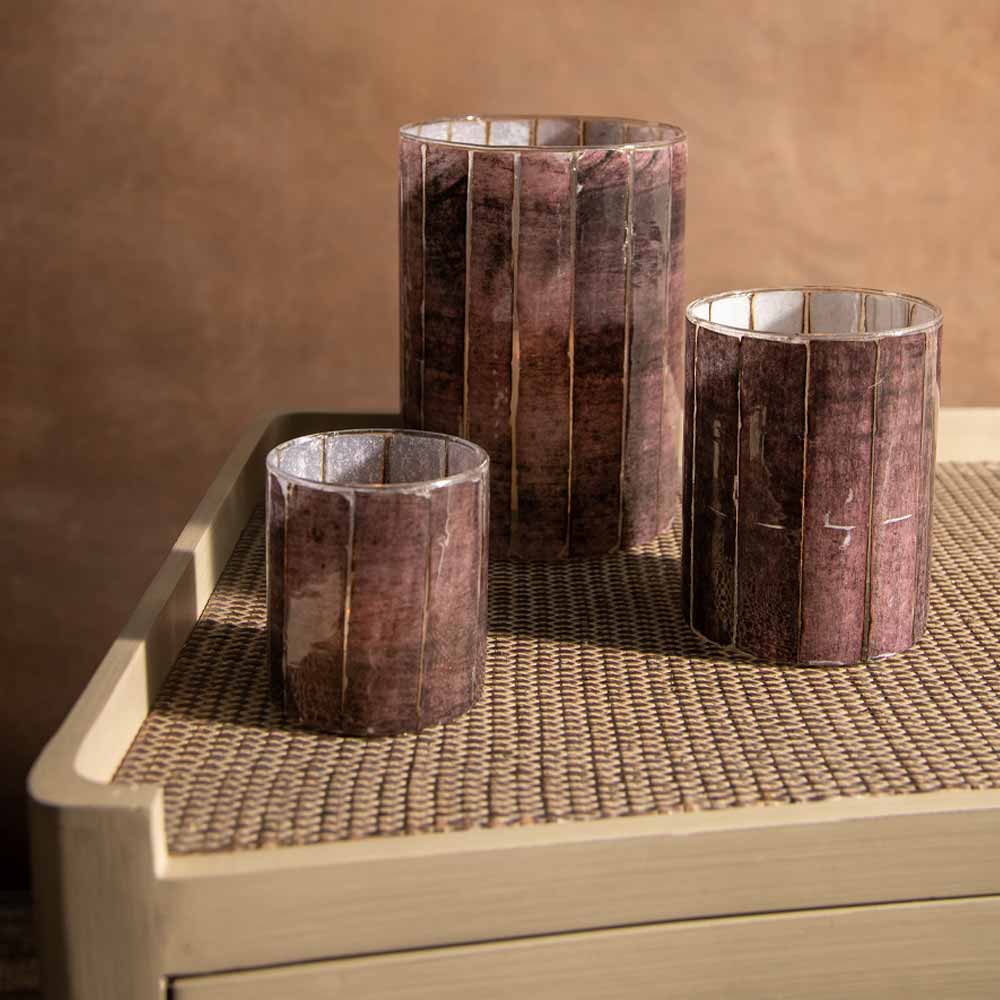Three burgundy striped glass candle holders with a textured,  arranged on a light wood side table against a warm brown background.
