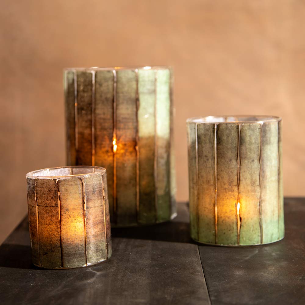 Three ribbed glass candle holders with a green-to-brown gradient finish, lit with warm candles, arranged on a dark tabletop against a soft brown background.