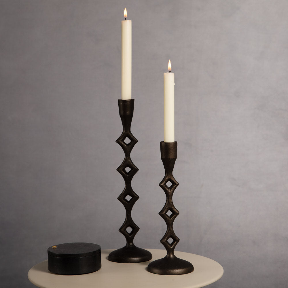 Two black metal candlestick holders with a diamond-cut pattern, each holding a lit cream dinnercandle, displayed on a round table against a textured gray background. A small round black box is also visible on the table.