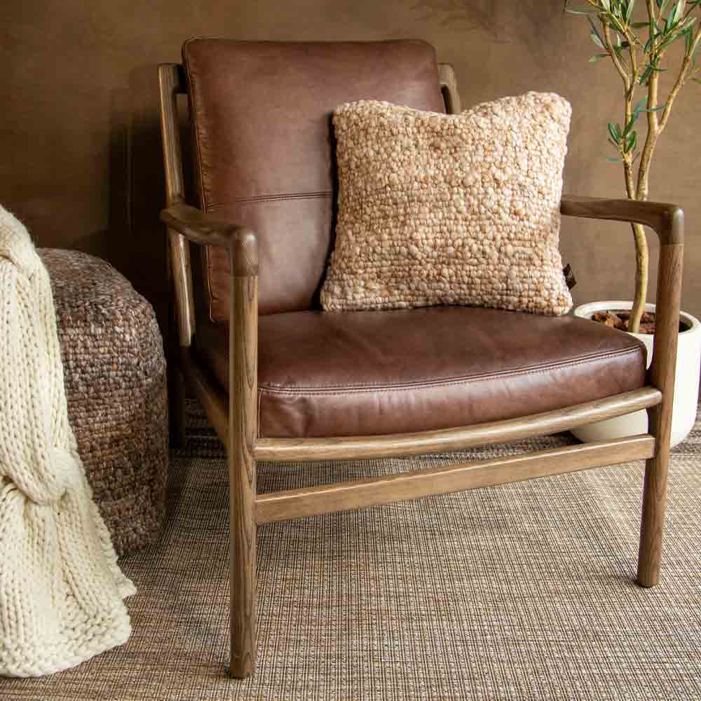 A mid-century modern wooden armchair with a brown leather seat and back, styled with a chunky beige textured  pillow, set on a woven rug against a brown wall.