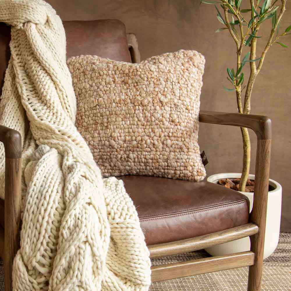 A square textured beige pillow styled on a brown leather armchair with a chunky cream cable-knit throw draped over the arm and a potted olive tree in the background.