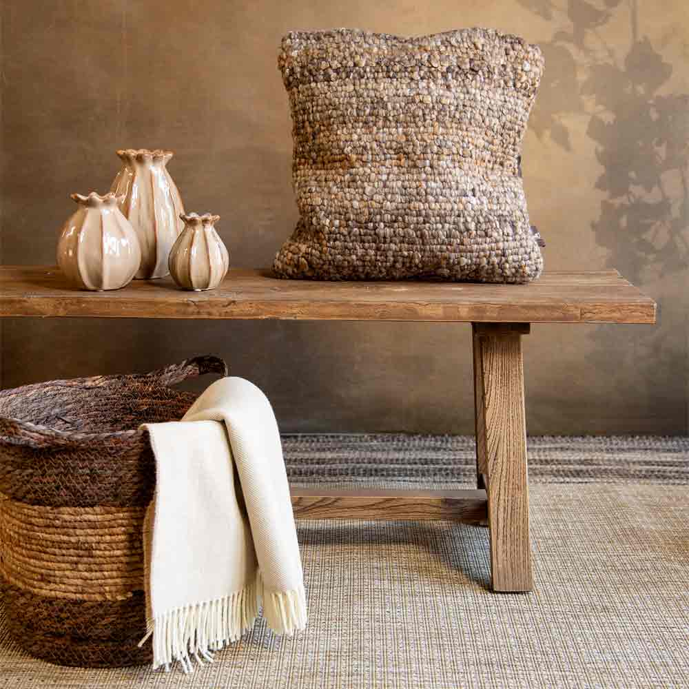 A square chunky woven cushion in a natural cream and beige fiber blend sits on a rustic wooden bench, styled with ceramic vases, a rope basket, and a cream throw blanket against a textured brown wall