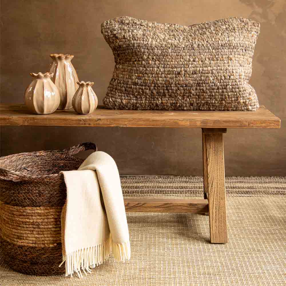A large rectangular chunky woven throw pillow in a natural beige and brown weave, styled on a rustic wooden bench next to a cream throw blanket and decorative ceramic vases.