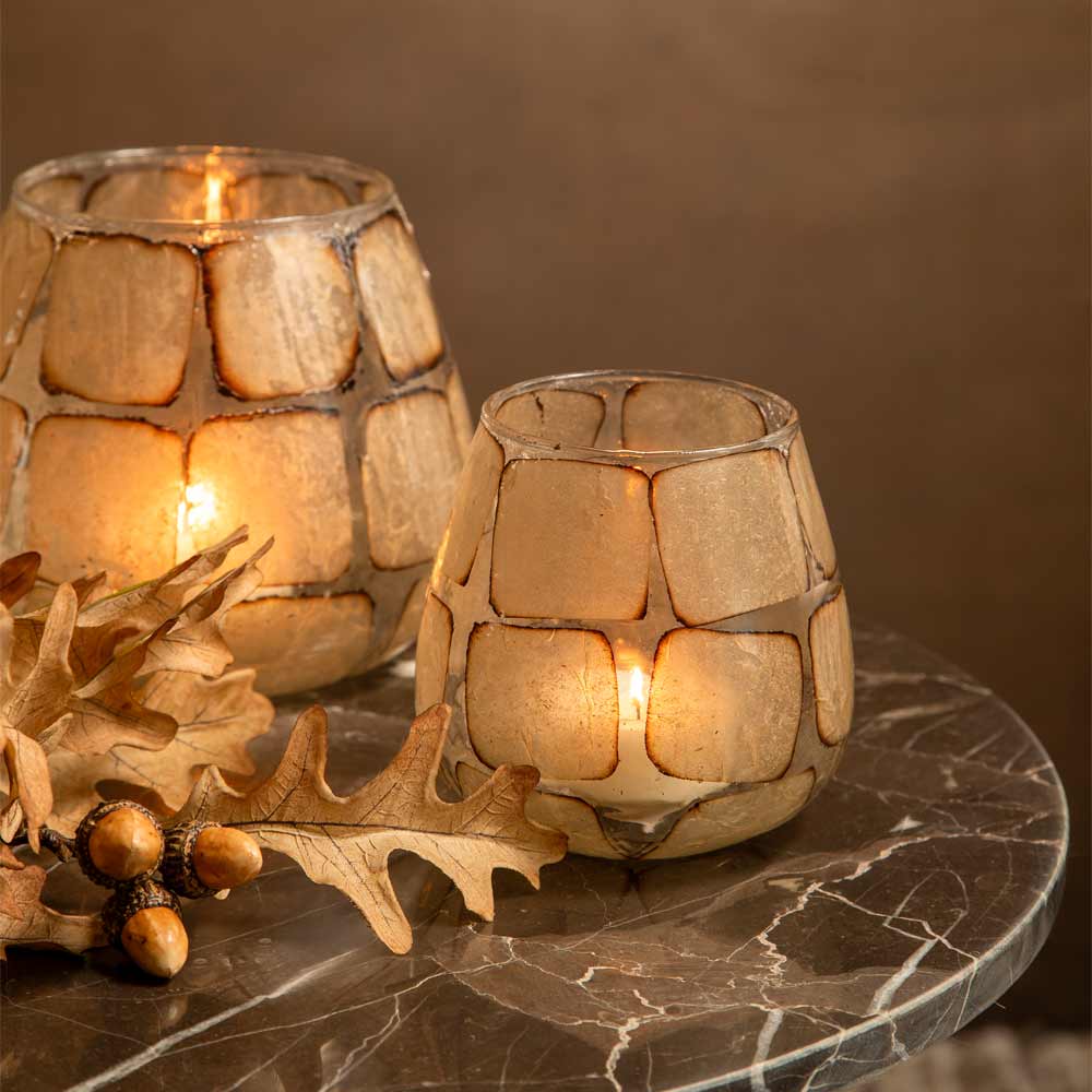 Two glass candle holders lit with warm Tealight candles, displayed on a dark marble surface with decorative oak leaves and acorns.