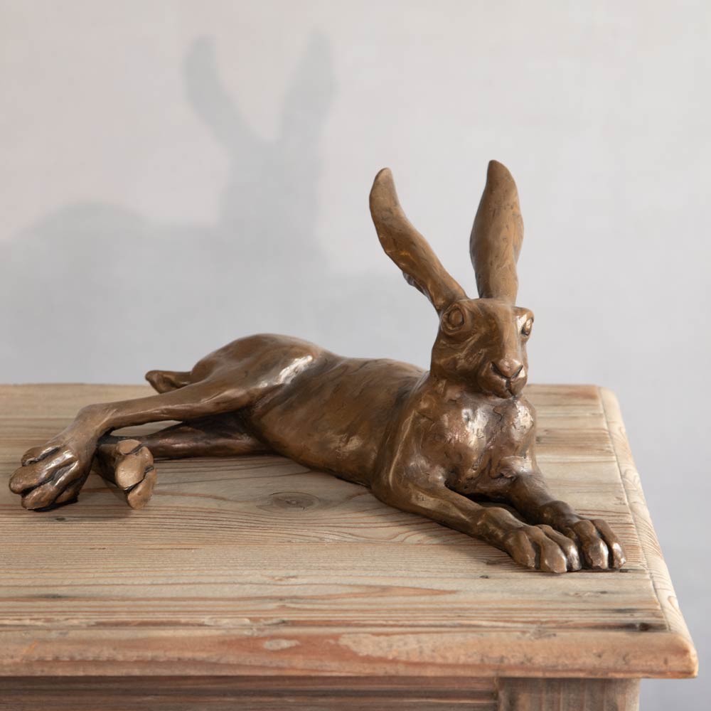 Bronze-finished sculpture of a hare lying down, displayed on a rustic wooden table against a light gray wall with a soft shadow cast behind it.