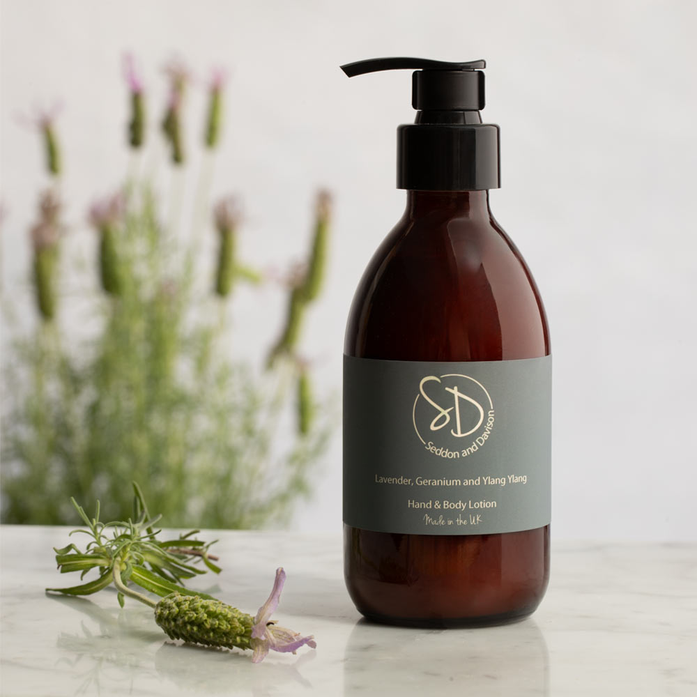Amber glass pump bottle of hand and body lotion with a black pump and a dark gray label, placed on a light marble surface with lavender sprigs and blurred lavender plants in the background.