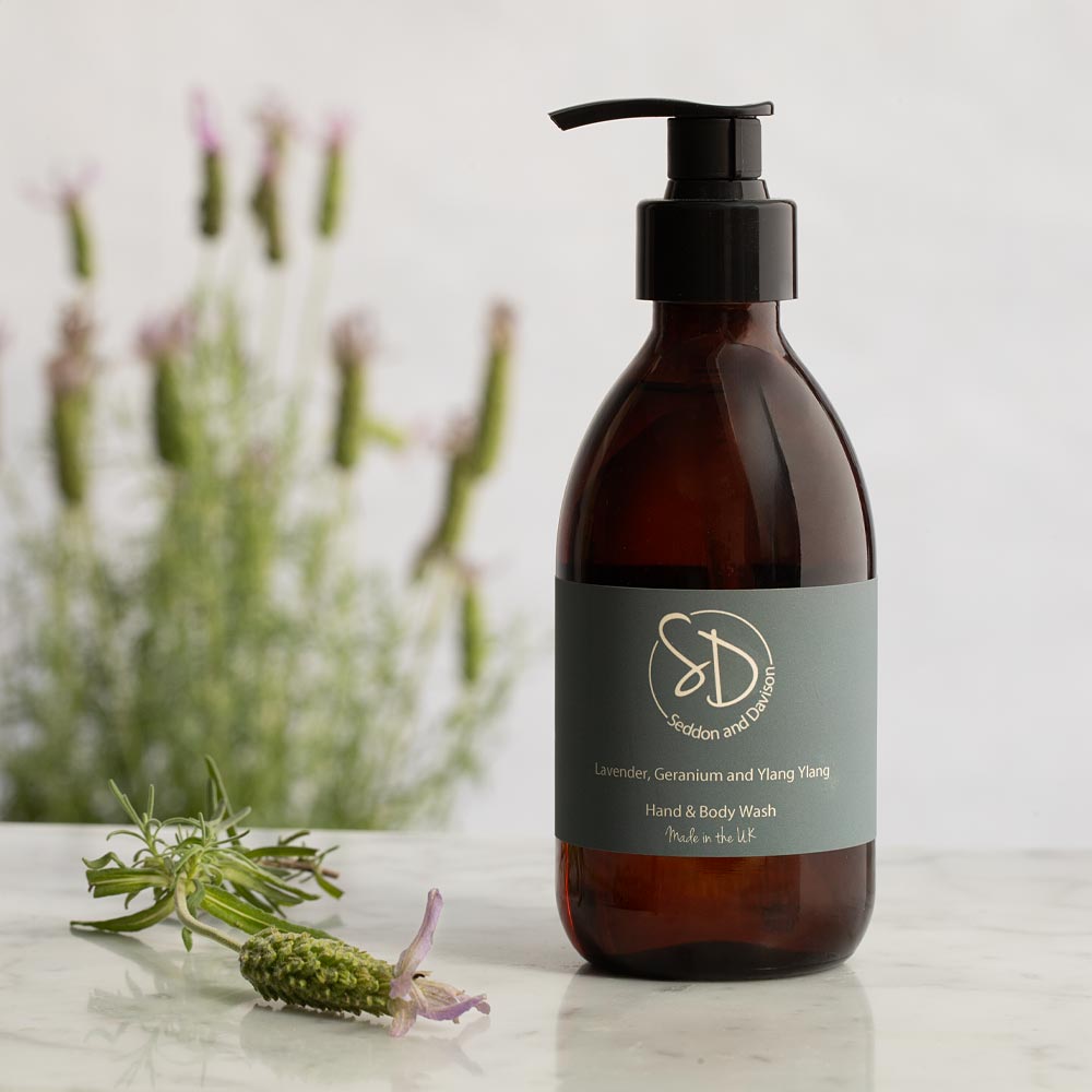 Amber glass pump bottle of hand and body wash with a black dispenser and a gray label, placed on a light marble surface with blurred lavender flowers in the background.
