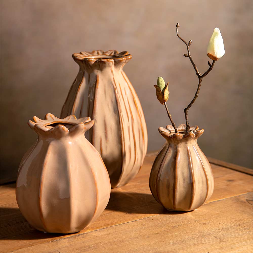Three ceramic bud vases in a warm taupe and brown glaze, arranged on a wooden surface against a textured neutral background. two vases are empty, while the other one hold a single decorative faux branch with small buds.