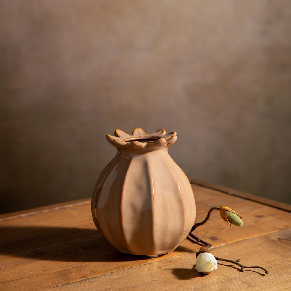 A small matte ceramic bud vase with a flower-pod silhouette sits on a rustic wooden table against a warm brown textured wall. A dried flower bud and white blossom rest beside it.