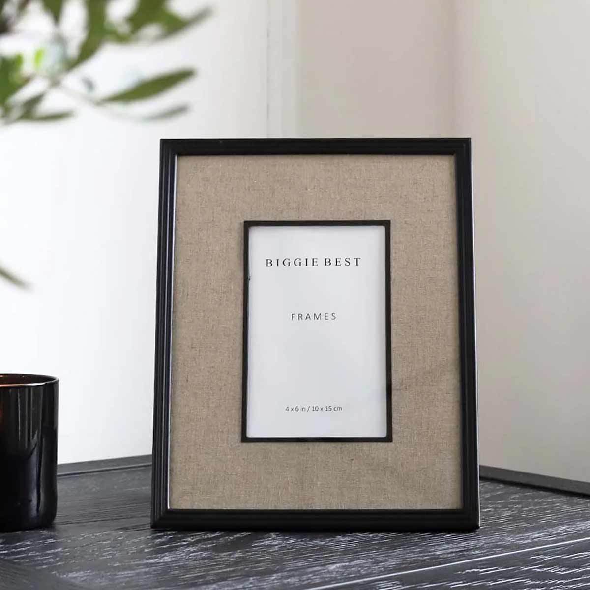 A black photo frame with a linen border displaying - 4×6 inch size. The frame is placed on a dark surface with a plant in the background.