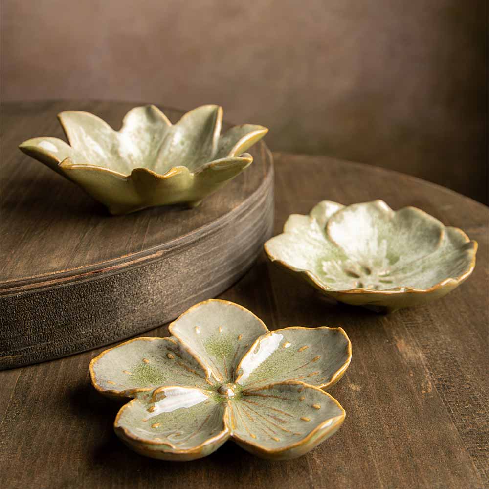 Three ceramic flower-shaped bowls with a glossy pale green glaze and gold-toned edging, arranged on a dark wooden surface with one bowl resting on a round wooden block.