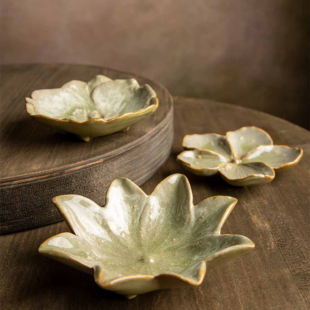 Three ceramic flower-shaped bowls with a glossy pale green glaze and gold-toned rims, arranged on a dark wooden surface with one bowl elevated on a round wooden riser.