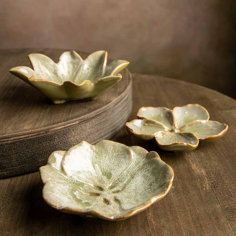 Three ceramic flower-shaped dishes with a glossy sage green glaze and gold-toned edges, arranged on a dark wooden surface.