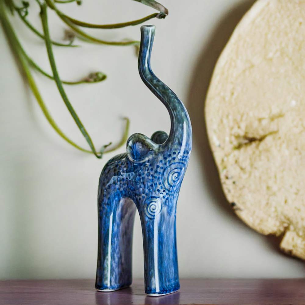 Blue reactive-glazed elephant ornament with wave patterns on a wooden surface with a plant and round object in the background