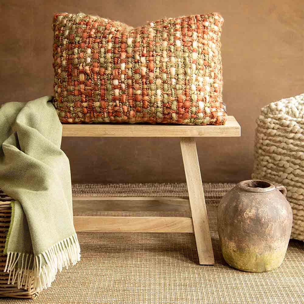 A rectangular woven throw pillow in a textured blend of rust, beige, and muted green, displayed on a small wooden bench in a rustic room setting.