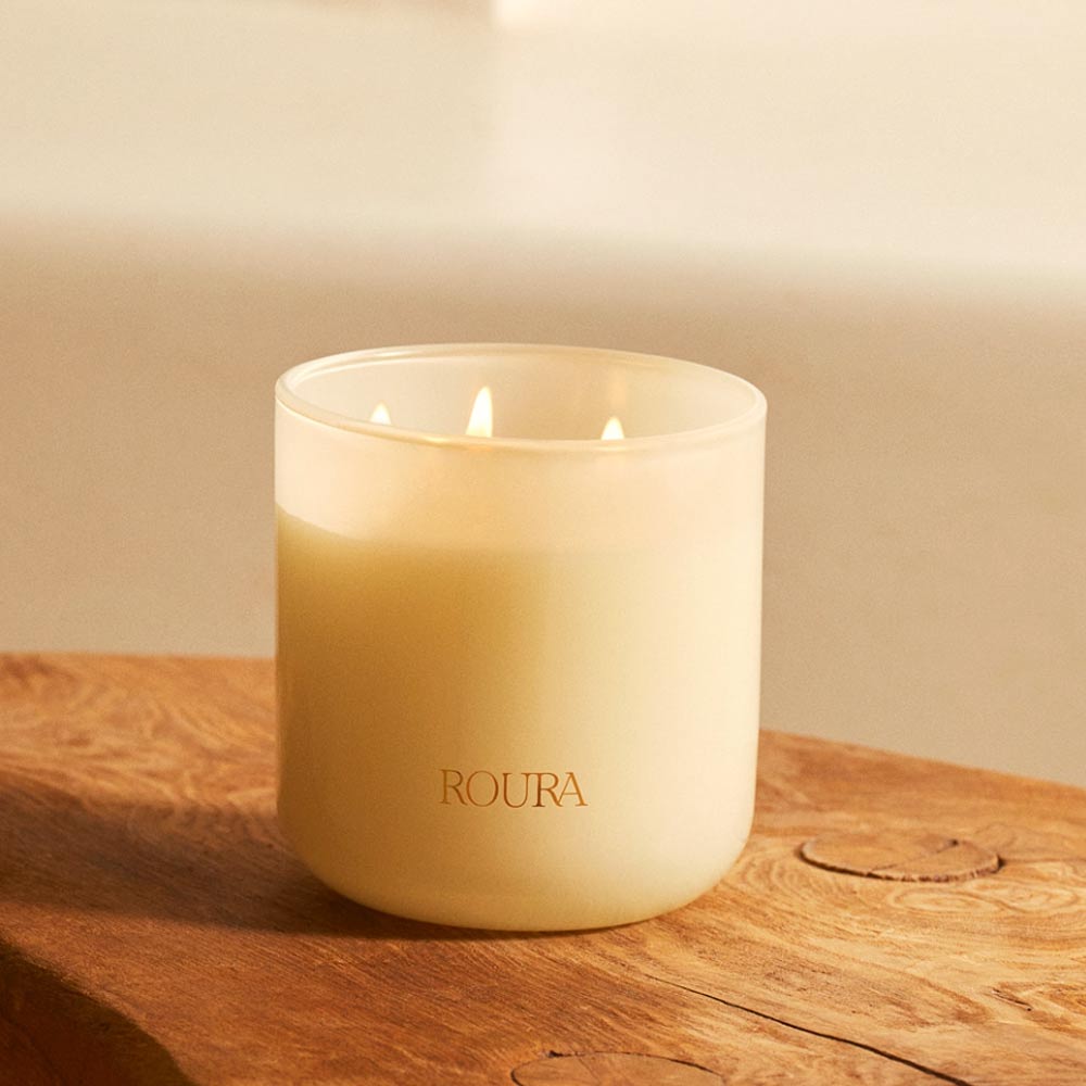 Lit Majestic Lotus 3 Wick Candle by Roura in a white glass container on a wooden surface.