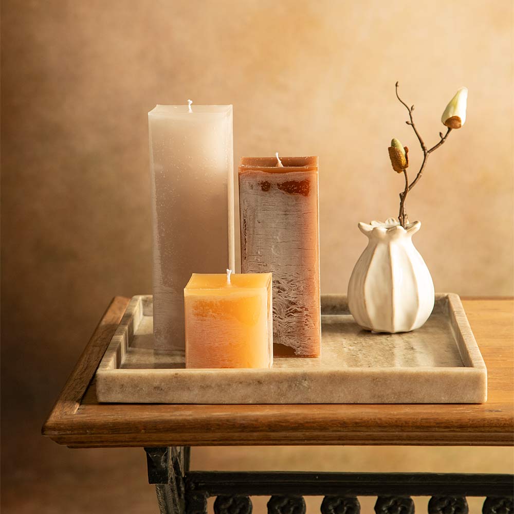 Three textured pillar candles in linen, caramel , and chocolate are arranged on a brown marble tray next to a small white vase with Faux magnolia  stem , set on a wooden table against a warm beige background.