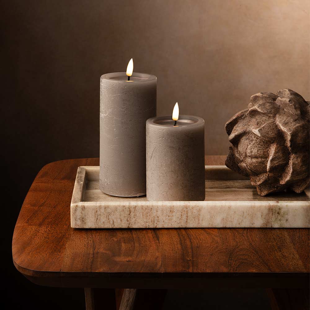 Two lit mocca LED pillar candles on a rectangular brown marble  tray, placed on a wooden table with a decorative sculptural object beside them against a warm brown background.