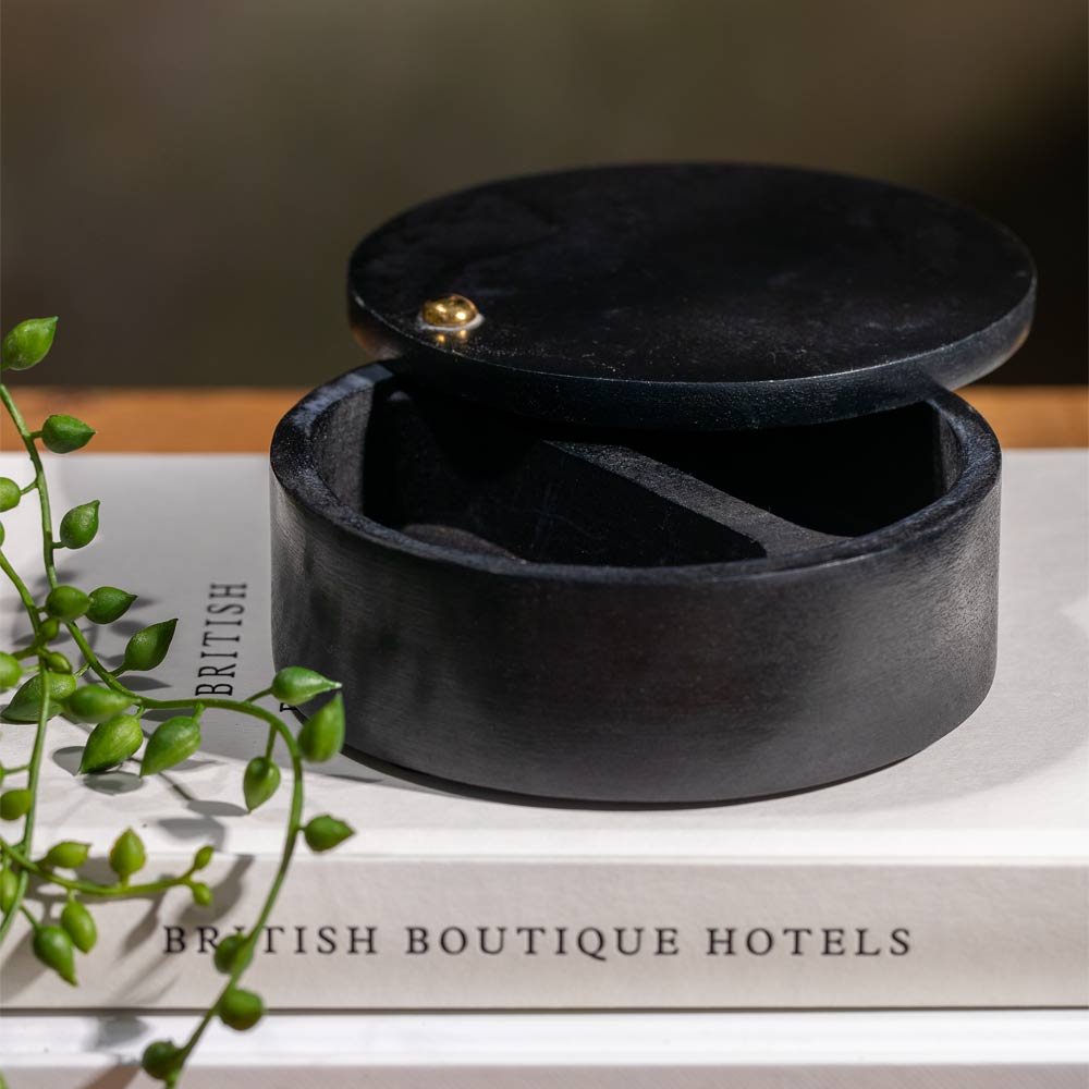 A round black wooden keepsake box with its lid slightly open, resting on a white book with green plant stems in the foreground.