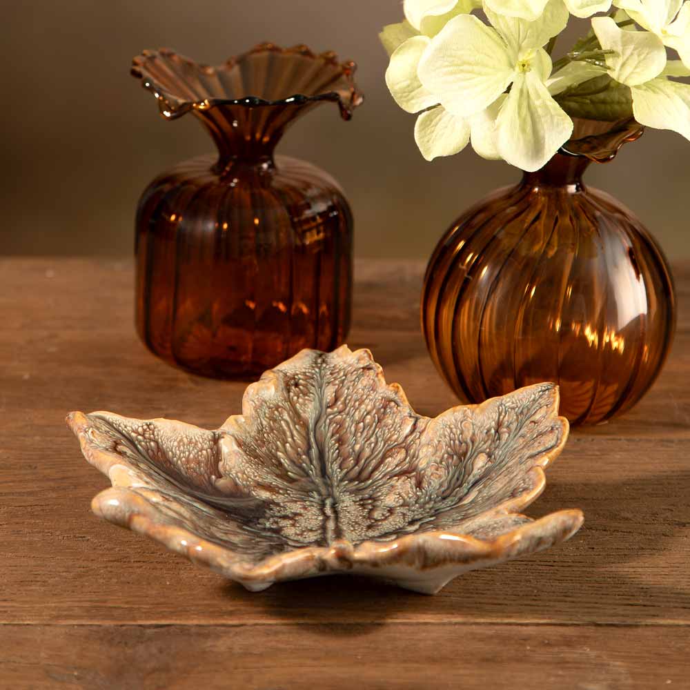 A ceramic maple leaf-shaped dish with a textured, marbled green amber  and beige pattern sits on a wooden table, with two amber glass bud vases one containing a sprig of green hydrangea flower in the background.