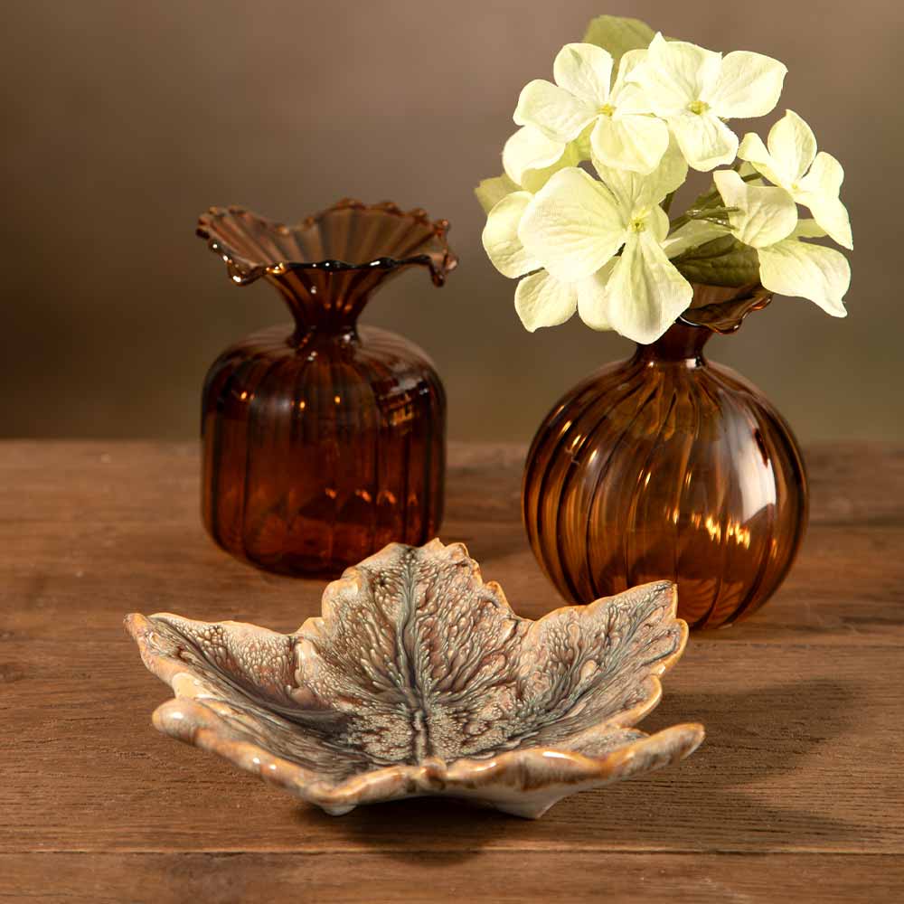 Two amber brown ribbed glass bud  vases with one holding a sprig of a pale green hydrangea flower, set on a wooden table next to a ceramic maple leaf-shaped dish.