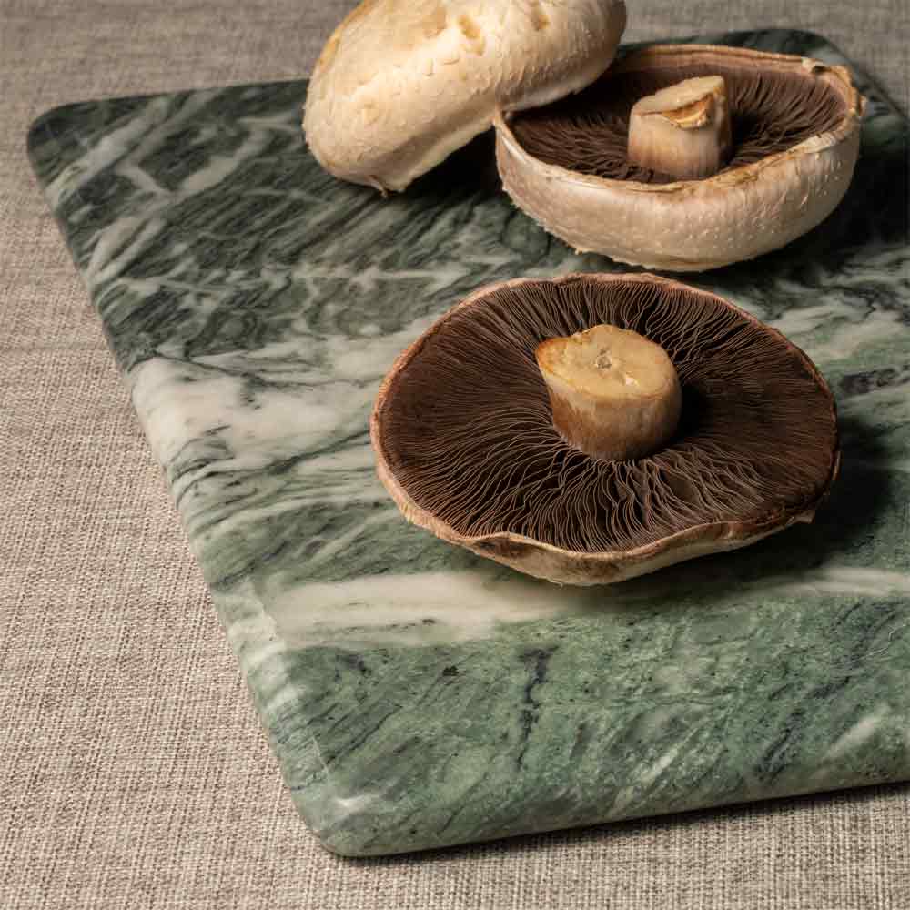A green and white veined marble cutting board with whole mushrooms placed on top, set on a textured beige linen surface.