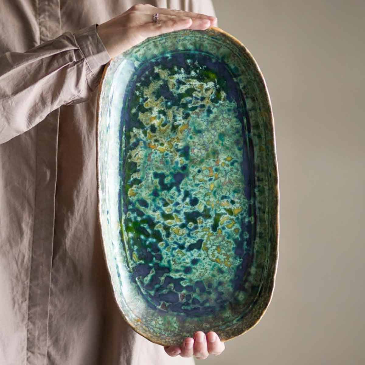 Hands holding the Milani Green Stoneware Serving Plate, showcasing its reactive glaze with deep green, ocean blue, and golden hues.
