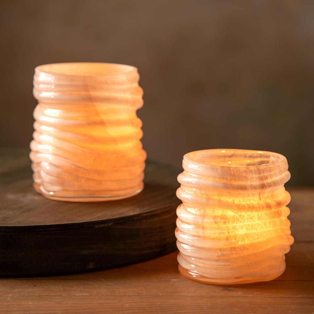 Two ribbed glass candle holders glowing with warm  light from a tea light candle, one standing on a dark wooden riser and the other on a wooden surface against a soft brown background.