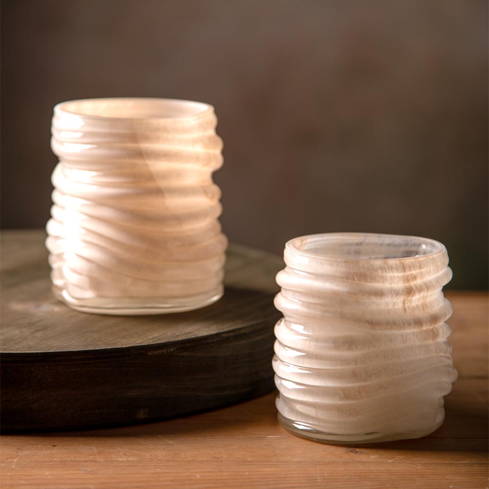Two small ribbed glass candle holders with a wavy, layered design, one resting on a dark wooden riser and the other on a wooden surface, set against a warm, dark brown background.