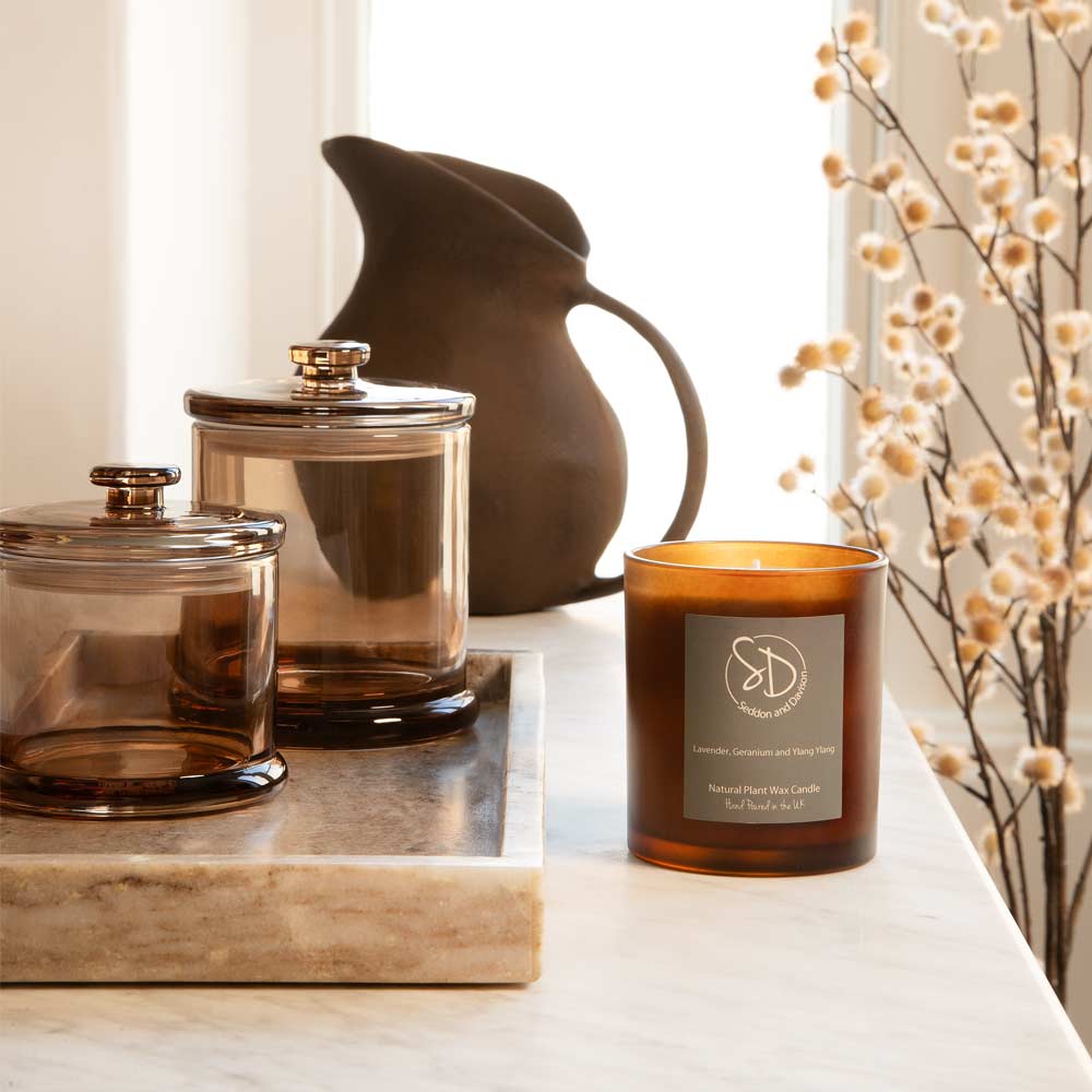 A lavender and geranium amber glass jar candle sits on a white surface beside a brown marble tray containing two amber glass jars with lids, in the background a dark ceramic pitcher and faux floral branches shield light streaming in from the window.