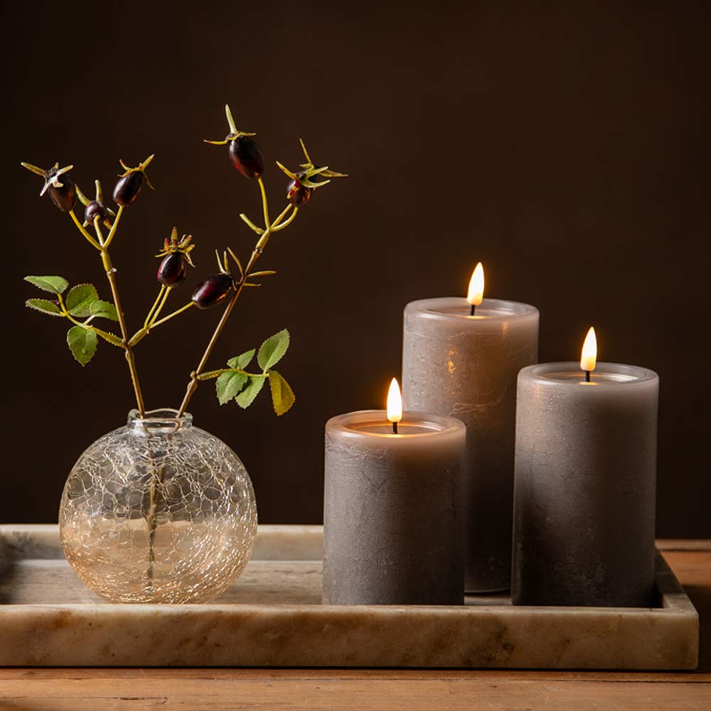 Three lit mocca pillar candles arranged on a marble tray beside a glass vase with faux  rose hips, against a dark brown background.