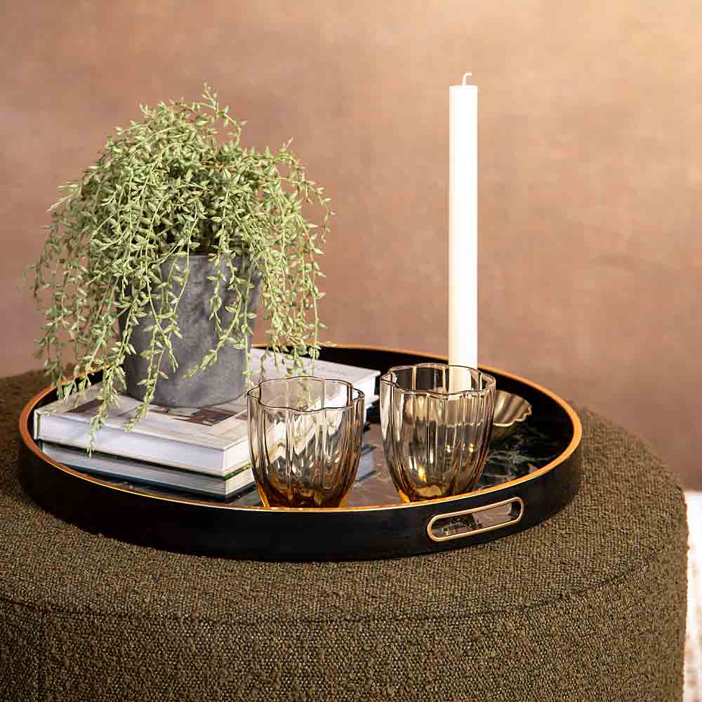 A round black serving tray with a gold-trimmed edge and a cutout handle, styled on a textured olive green ottoman. The tray holds a potted trailing green plant, a stack of books, two amber-tinted glass tumblers, and a tall white candle against a warm brown wall background.