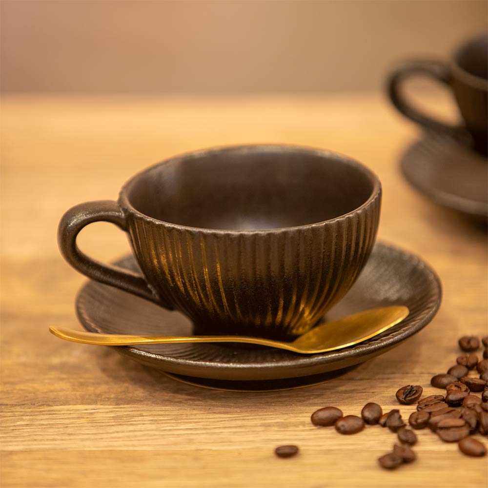 Black ceramic coffee cup and saucer with a gold spoon on a wooden table, surrounded by coffee beans.