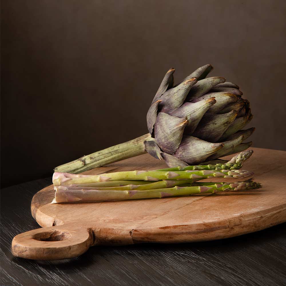 A whole artichoke with green stalk and several asparagus spears arranged on a round wooden cutting board against a dark brown  background.