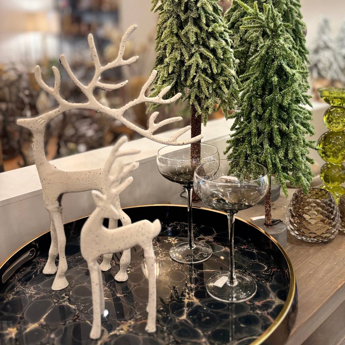 White and grey slim Nordic Olaf stag ornaments styled with wine glasses on a marble tray with festive trees