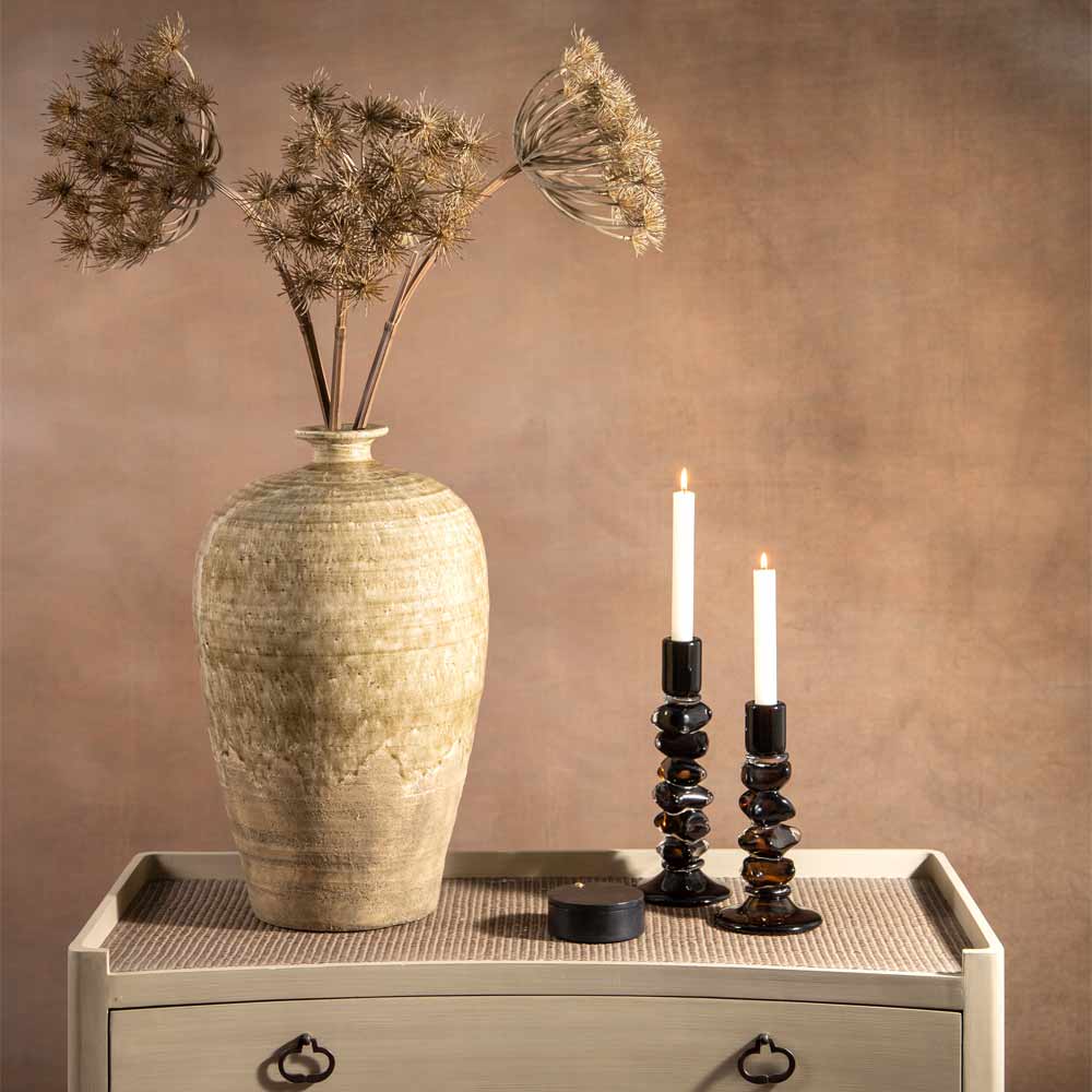 A rustic olive glazed ceramic vase with  faux Queen annes lacec stems sits on a light wood console table against a warm brown textured wall. Two dark olive candlestick holders with lit white candles and a small black round lidded jar  are placed beside the vase.