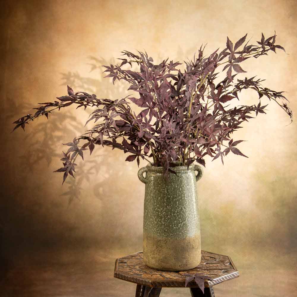 A terracotta vase with a speckled green and beige glaze, holding faux Acer branches with dark burgundy foliage, displayed on a small wooden table against a warm, textured brown background.