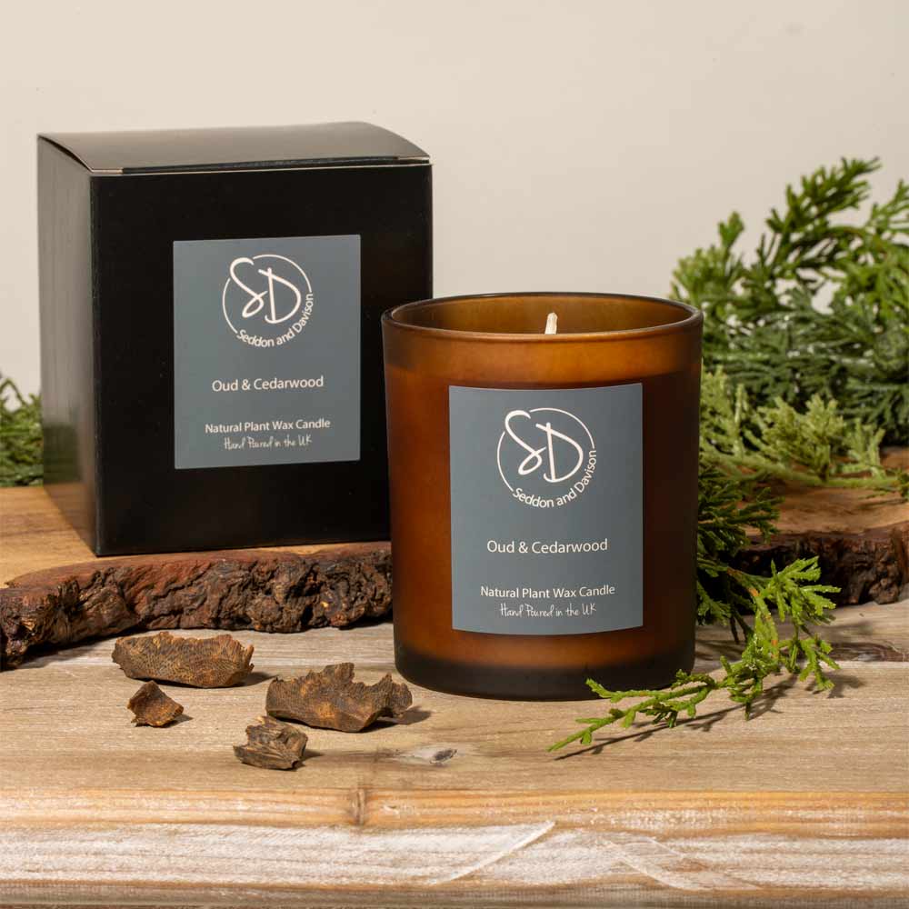 An amber glass jar candle labeled 'Oud & Cedarwood' stands on a rustic wooden surface beside its black product box. Greenery and bark props are arranged around the candle, with a soft neutral wall in the background.