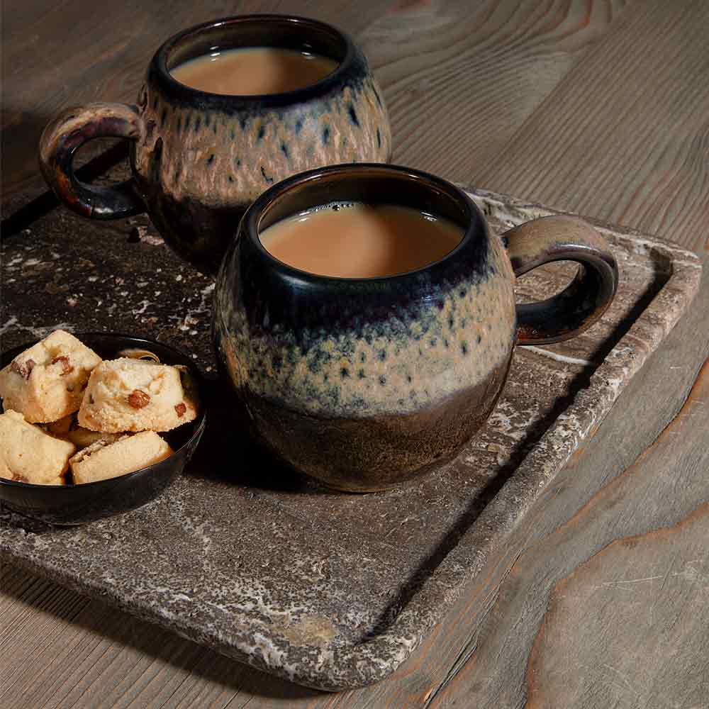 Two ceramic mugs filled with Tea  arranged on a rustic stone tray with a small bowl of cookies on a wooden table.