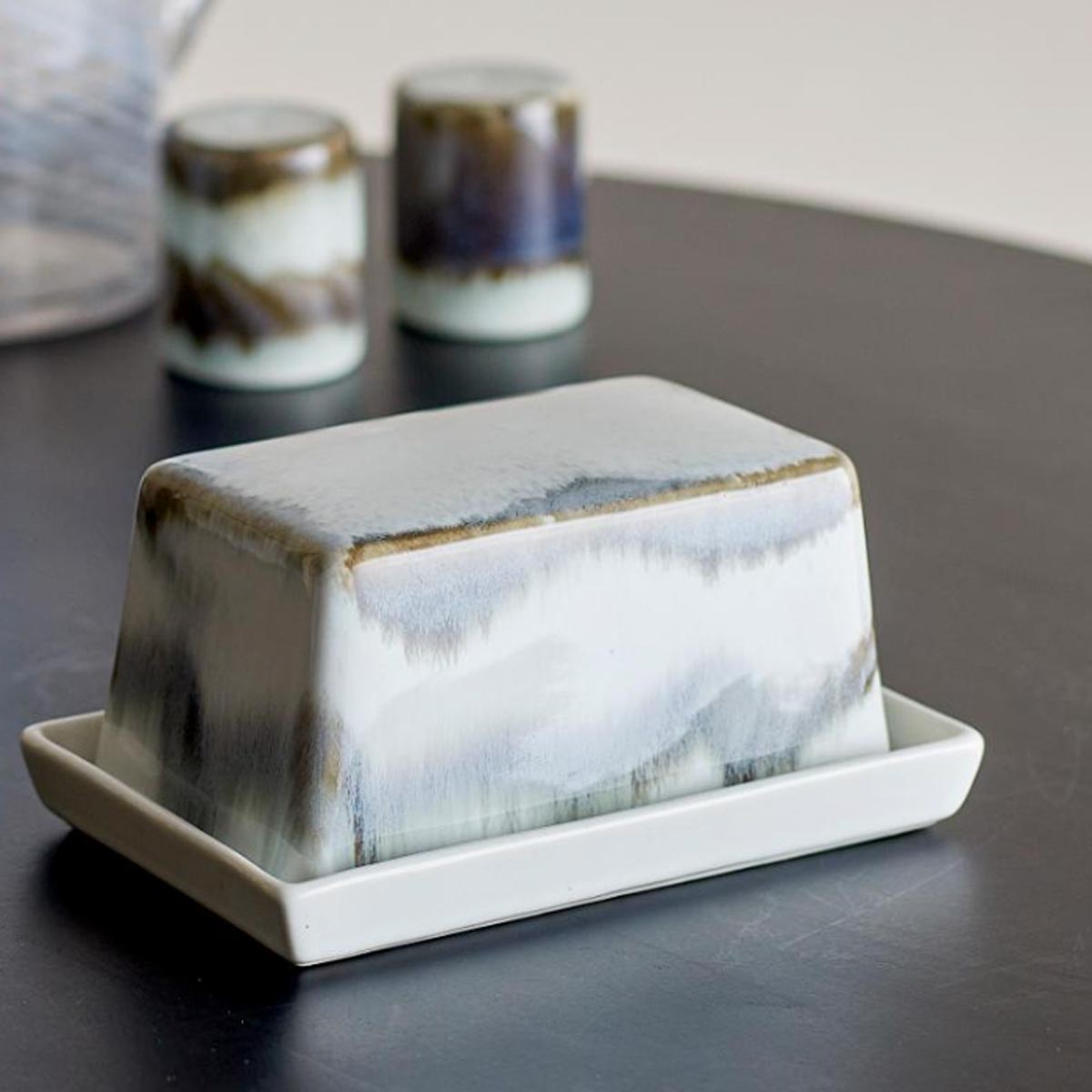 Paula Stoneware Butter Dish with a reactive glaze in grey and white, placed on a black table with salt and pepper shakers in the background.