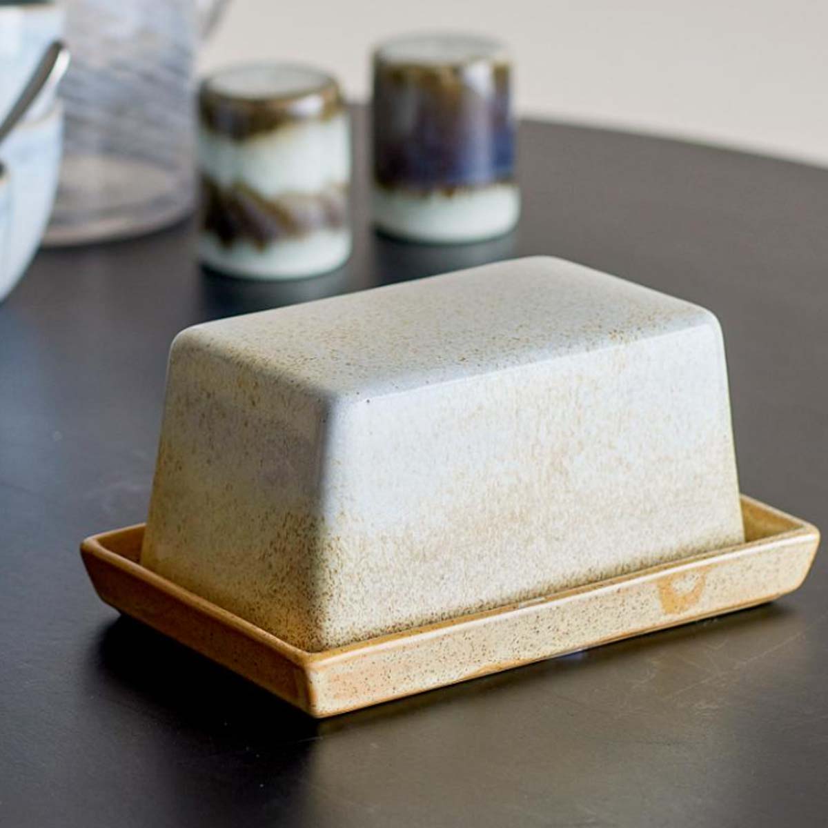 Paula Stoneware Butter Dish in natural golden hues, placed on a black table with salt and pepper shakers in the background.