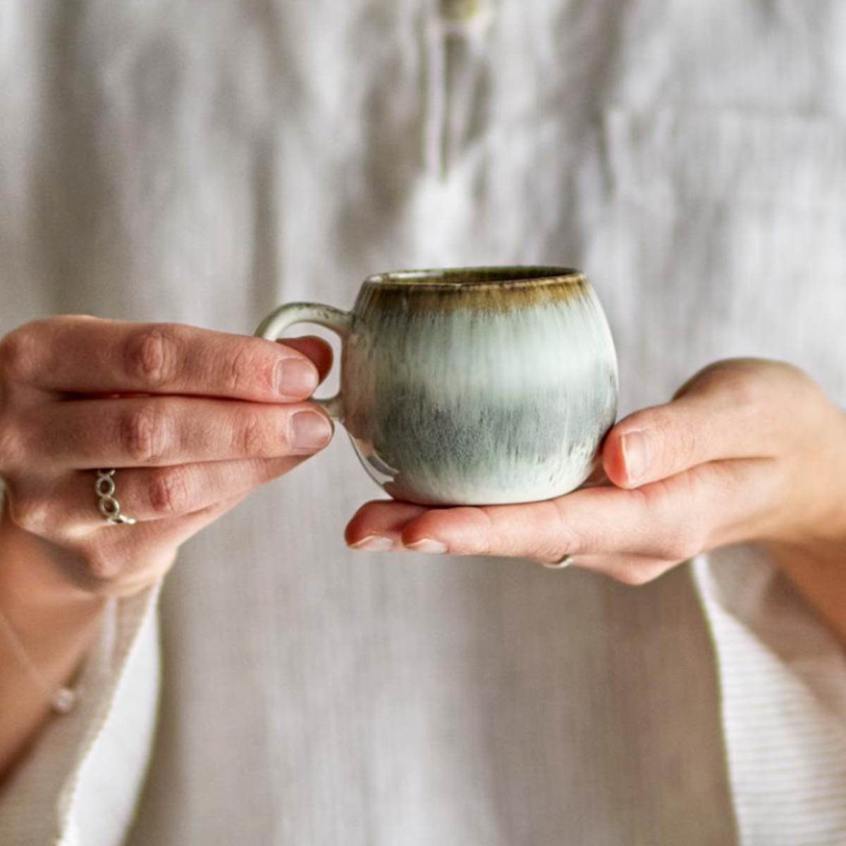 Paula Stoneware Espresso Cup in grey, white, and brown reactive glaze, held in both hands, showcasing its organic shape and unique colour variations.