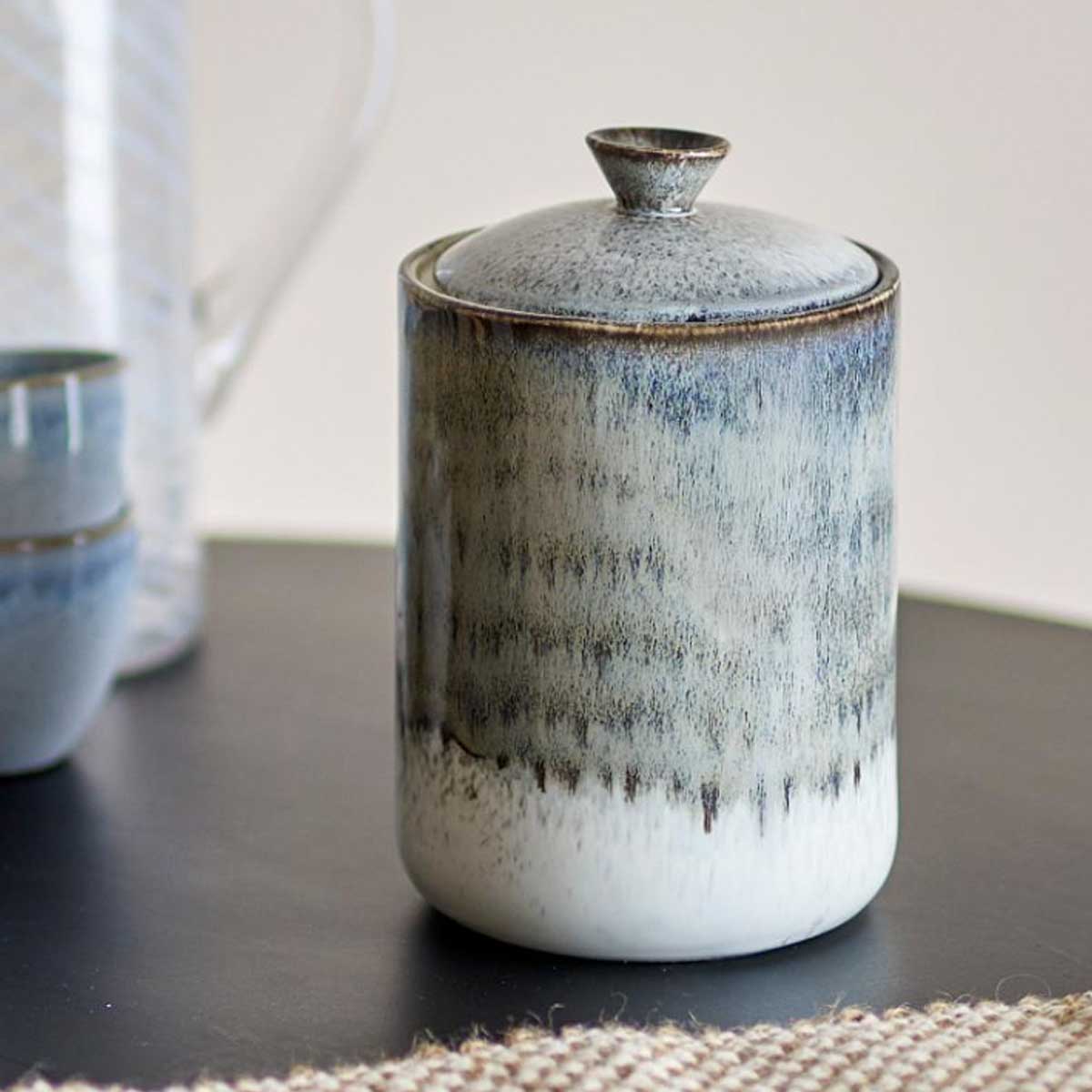 Paula Stoneware Jar in grey with a reactive glaze, featuring a lid with a silicone seal. Displayed on a dark surface with ceramic bowls and a glass jug in the background.