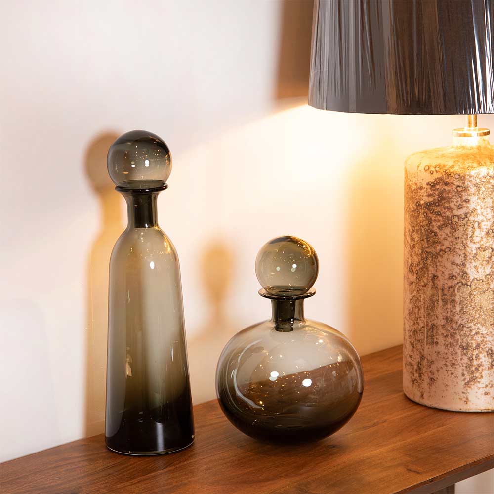 Two Pearl Grey Glass Bottles with Ball Tops on a wooden surface with a lamp in the background