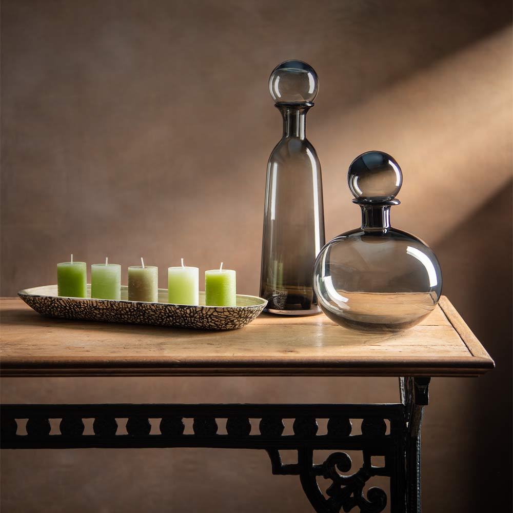Two dark glass decanter style vases with spherical stoppers and five green pillar candles on a speckled tray, displayed on a wooden table against a warm brown background.