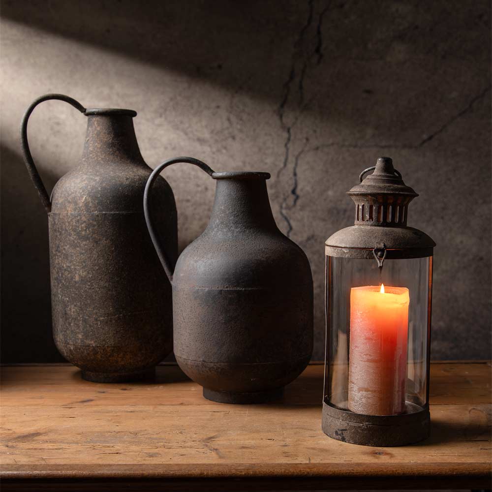Two rustic dark metal vases with handles sit on a wooden table beside a matching lantern-style candle holder with a lit pillar candle, against a textured old wall with dramatic shadows.
