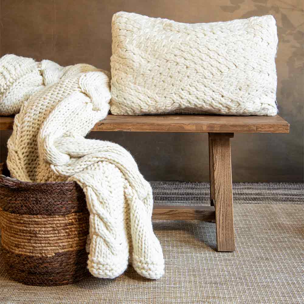 A chunky knit cream throw blanket and matching textured cream pillow displayed on a rustic wooden bench over a woven rug.