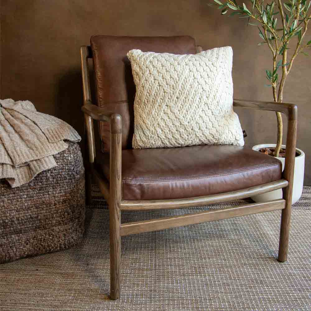 A mid-century modern armchair with a brown leather seat and backrest, a light wood frame, and a cream textured knit throw pillow, styled in a rustic room setting.