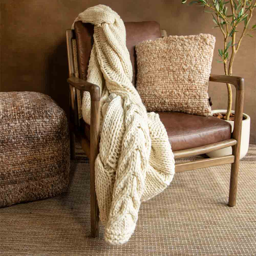 A chunky cream knit throw blanket is draped over a mid-century modern wooden armchair with brown leather upholstery. The chair is styled in a cozy room setting with a textured beige throw pillow, a brown woven pouf, and a potted plant.