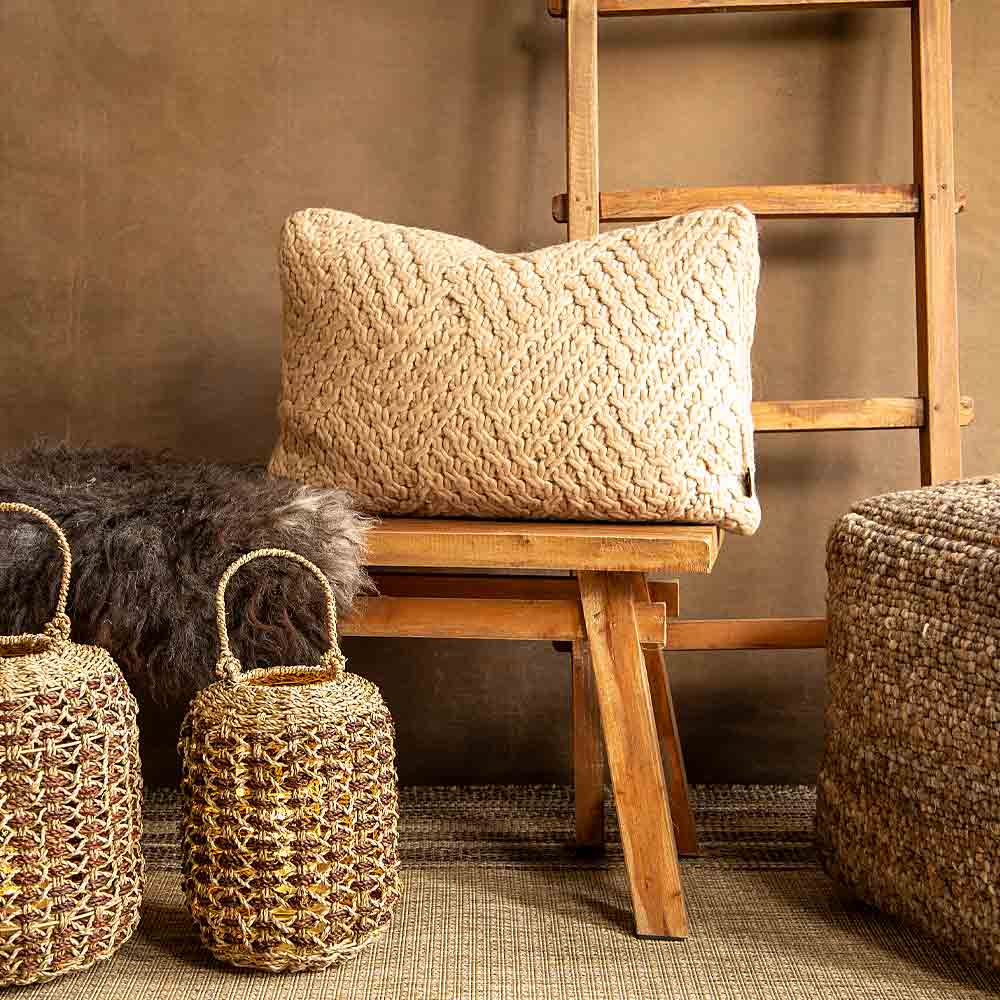 A rectangular woven chunky Knit  pillow in a natural Taupe colour, styled on a wooden bench  in a rustic room setting with a wooden ladder, woven lanterns , and a dark sheepskin rug.